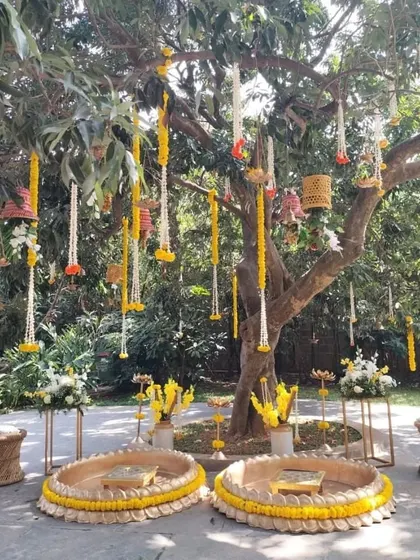 A stunning Haldi ceremony setup under a tree, decorated with hanging marigold and tuberose garlands. The low, circular seating arrangements are perfect for the rituals and for guests to gather around.