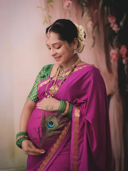 An elegant and traditional maternity portrait. The client is dressed in a beautiful pink and green saree, adorned with classic jewelry and a peacock feather for a culturally rich image.