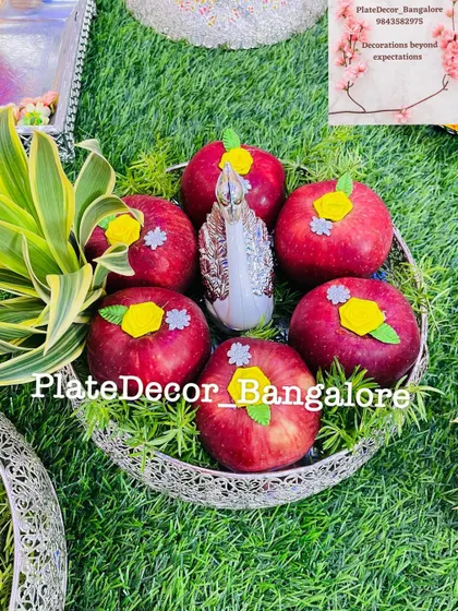 A silver bowl of red apples, each decorated with a yellow flower and a silver star, with a swan figurine in the center.