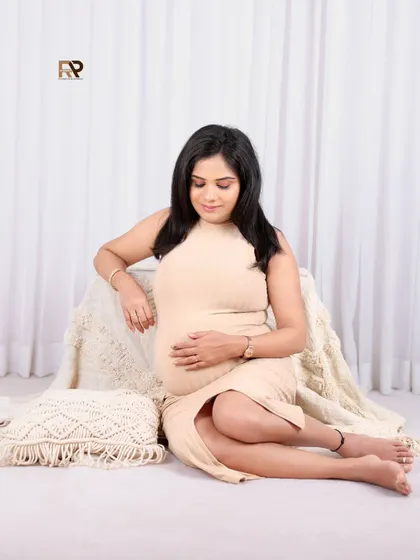 A peaceful moment of reflection. The mom-to-be is seated in a bohemian-inspired setup with macrame pillows, her simple beige dress creating a natural and earthy vibe.