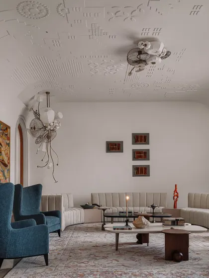 The formal living room at Rehethaan, where high-backed blue armchairs and a channel-tufted sofa create an elegant seating arrangement. The space is unified by a large, patterned rug and adorned with sculptural lighting.