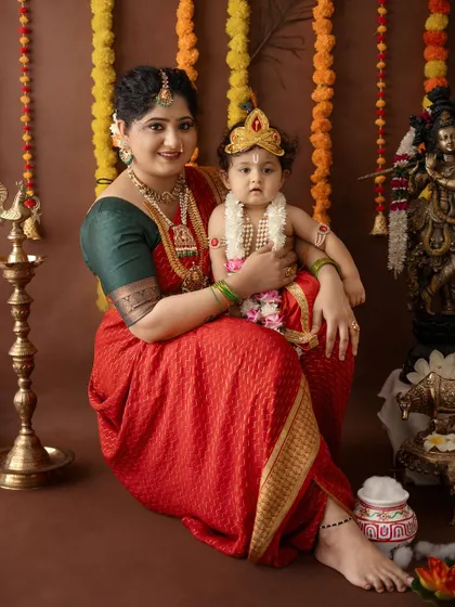 A serene portrait of a mother and her baby dressed as Krishna, surrounded by traditional brass lamps and flower garlands.