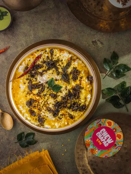 A top-down photograph of tempered curd rice, styled to feel comforting and soulful. The dark, moody background makes the vibrant yellow tempering with curry leaves and red chili pop, telling a story of simple, homemade goodness.