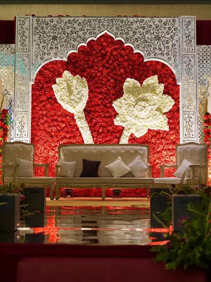 A stunning stage backdrop for a 'Ruby Romance' themed wedding. A wall of red roses is accented with white floral lotuses and framed by intricate, traditional white panels.