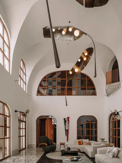 A wide shot of the double-height living room at Rehethaan, showcasing the grotto-like architecture, arched windows, and the magnificent leaf-like light installation that hangs from the ceiling.