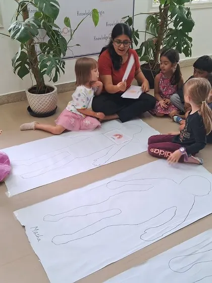 A teacher discusses the different parts of the human body with a group of students, using their body outlines as a visual aid.