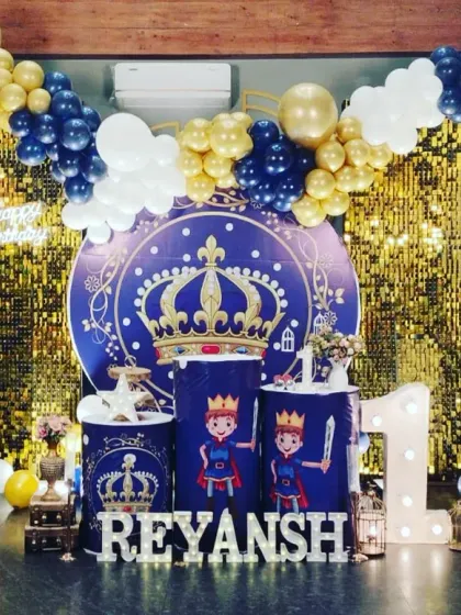 A royal celebration for Prince Reyansh's first birthday. This grand setup features a gold sequin backdrop, a royal blue and gold balloon arch, and a crown-themed backdrop for the little prince.