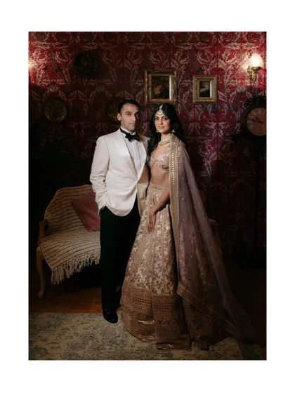 A vintage-inspired couple portrait against a rich, patterned wallpaper. The groom's classic tuxedo and the bride's Sabyasachi lehenga create a look of old-world glamour.