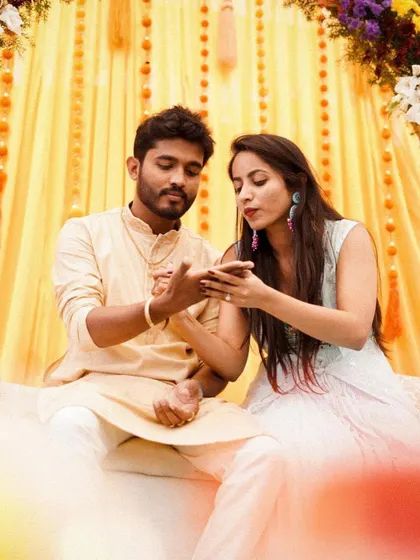 A sweet moment from a Mehendi ceremony. The decor features a bright yellow backdrop with colorful floral arches, creating a cheerful and festive atmosphere.