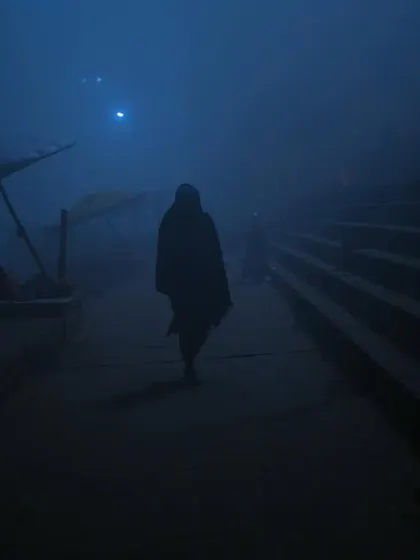 A lone figure walks along the ghats in the deep blue fog of pre-dawn Varanasi. The silhouette creates a mysterious and contemplative mood, reflecting the city's timeless nature.
