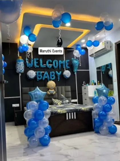 A 'Welcome Baby' room decoration in blue and white. The setup includes 'Welcome Baby' foil letters over the bed, balloon columns, and balloons on the ceiling.