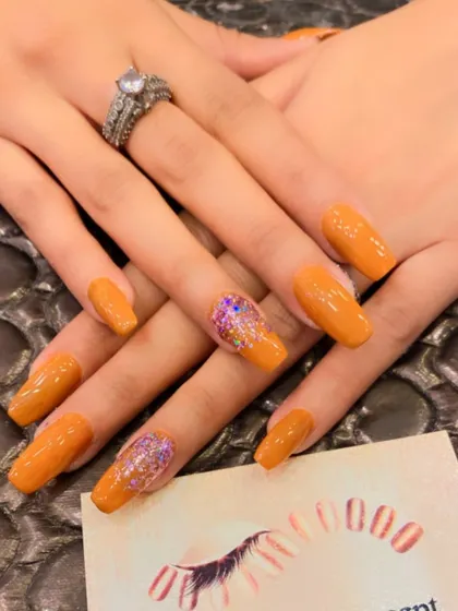 A vibrant orange set of coffin nails. The ring fingers are accented with a beautiful cascade of multi-colored glitter for a fun, sparkling contrast.