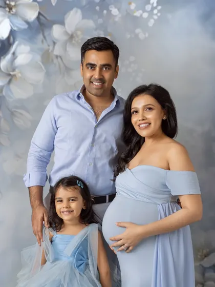 A bright and happy family portrait. The family smiles warmly at the camera, their light blue outfits perfectly complementing the soft, painted background of their maternity photoshoot.