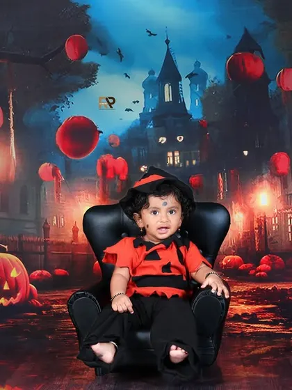 A baby dressed in a Halloween costume sits in a chair, looking like the cutest little pumpkin in the patch. The spooky backdrop with jack-o'-lanterns adds to the festive fun.