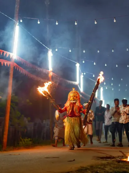 A full-length view of a Bhoota Kola performer, their traditional costume and flaming torches creating a striking silhouette against the night sky. This image captures the grandeur and mystique of the ceremony.