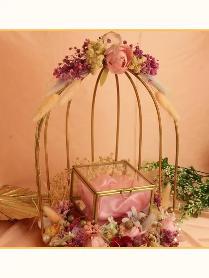 A beautiful engagement ring platter featuring a glass box nestled inside a decorative golden birdcage with floral accents. This design adds a touch of whimsy and elegance to the ceremony.