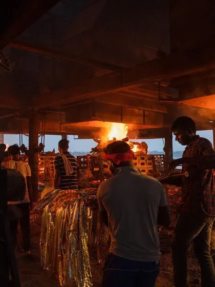 Workers tend to bodies wrapped in golden shrouds as pyres burn inside the crematorium. The image captures the ongoing, methodical nature of the work at the ghat.