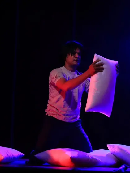 A solo performer kneels amidst a sea of pillows in "ME & MY PILLOW." This shot captures a moment of vulnerability and reflection within the larger contemporary dance piece.