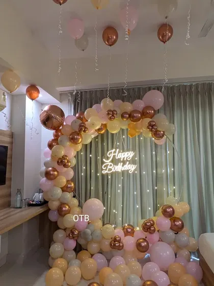 A cozy and elegant room setup featuring a circular balloon arch with a neon sign and fairy lights. The pastel and rose gold balloons create a warm and inviting atmosphere.
