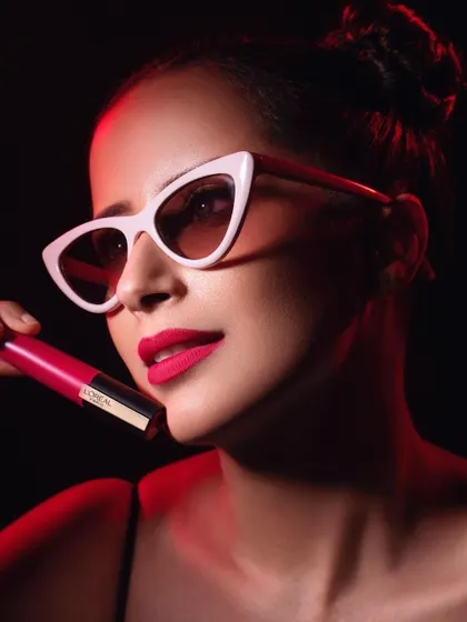 A chic and modern beauty shot for L'Oréal Lipstick Day. The model wears stylish cat-eye sunglasses and a bold pink lip, holding the matching lipstick. The red backlighting adds a dramatic, high-fashion flair, making the product feel both trendy and timeless.