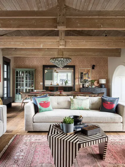 An open-plan living space in the Saratoga home, where the living room flows into the dining area. The design uses a consistent palette of warm woods and neutral tones, punctuated by colorful textiles and a striped coffee table. The exposed beam ceiling ties the entire area together, creating a cohesive and inviting environment.