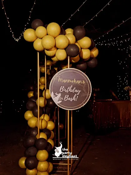 A custom circular sign for 'Manmohan's Birthday Bash' was integrated into a black and gold balloon column, serving as a stylish welcome marker for the event.