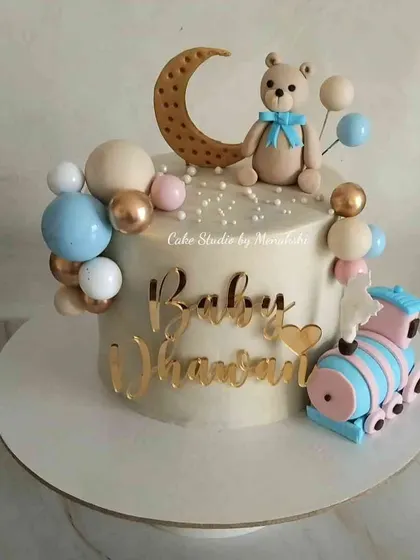A cute first birthday cake for baby Dhanvan. The design features a teddy bear on the moon, a little train, and balloons in soft pastel colors.