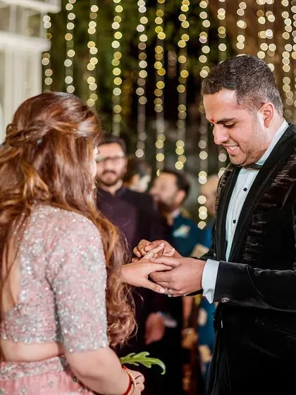 The groom, smiling, finally places the ring on his bride's finger, with family looking on.