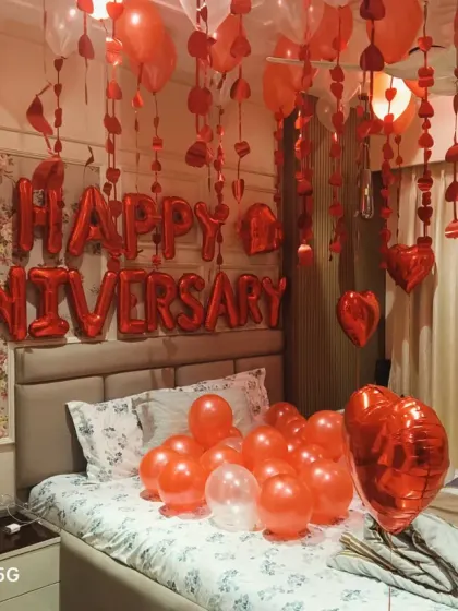 A full view of a romantic anniversary bedroom decoration. The room is filled with red heart-shaped balloons, hanging streamers, and large "Happy Anniversary" foil balloons above the bed.