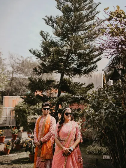 A wide shot of the couple posing in a garden during their Mehendi event, showcasing their coordinated outfits and the beautiful, natural setting.