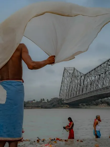 A man dries a large white cloth on the banks of the Hooghly river, with the Howrah Bridge in the background. The flowing cloth creates a sense of movement and freedom.