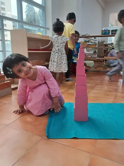 A young girl proudly stands next to the Pink Tower she has built. The sense of accomplishment that comes from completing a challenging task builds intrinsic motivation and self-confidence.