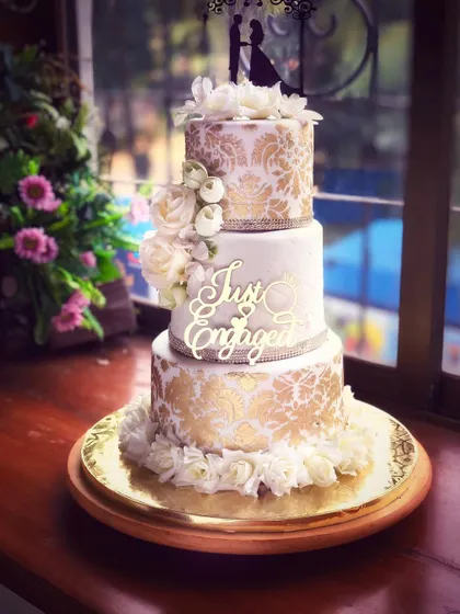 The stunning gold and white engagement cake displayed in natural light, showcasing its height and beautiful floral arrangement.
