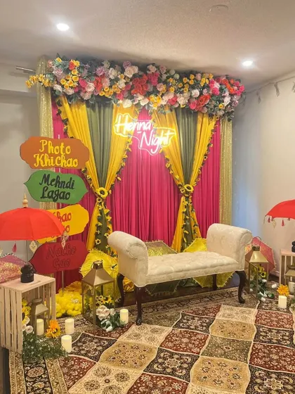 A fun and festive 'Henna Night' photo corner. The backdrop of pink and yellow drapes is accented with a floral top border, a neon sign, and quirky signboards for guests to pose with.