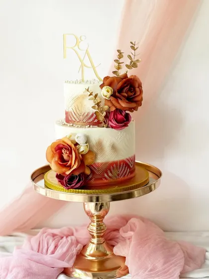 A two-tier cake with a beautiful burnt orange and red color scheme. The design includes intricate stencil work, gold accents, and is topped with silk flowers and a custom initial topper.