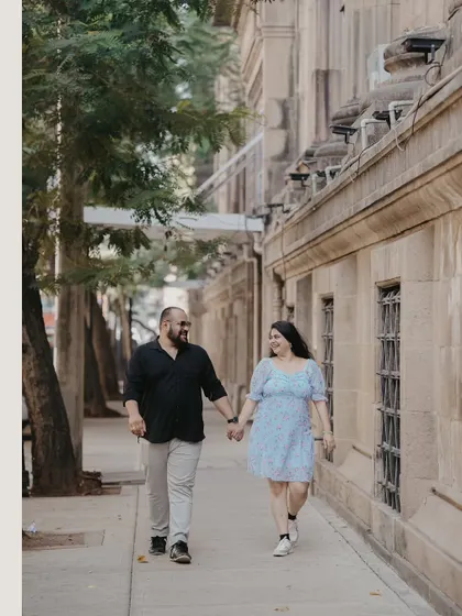 A couple walks hand-in-hand down a city sidewalk lined with trees and historic buildings. This classic walking shot captures a simple, romantic moment in the heart of the city.