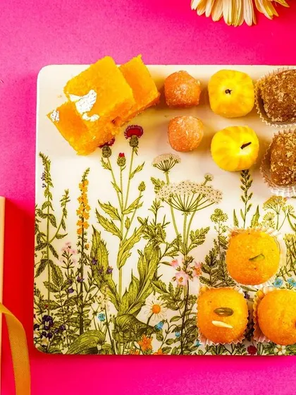 A flatlay of a serving plate with Indian sweets (mithai), styled with a gift box and flowers. This is a perfect shot for festive or wedding-themed content.