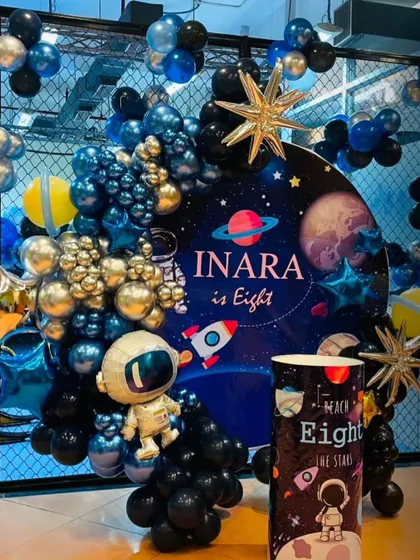 An elaborate space-themed party for an eighth birthday. This setup includes a detailed backdrop, an astronaut piñata, and a balloon arch in metallic blue, black, and gold.