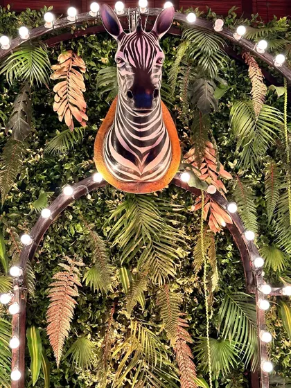 A zebra head sculpture mounted on a wall of lush greenery and framed by an arch of lights, a bold statement piece for a wild-themed event.