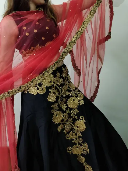 A detailed shot of the black lehenga, focusing on the heavy gold floral embroidery that climbs up the side of the skirt. The contrasting red dupatta adds a pop of color.