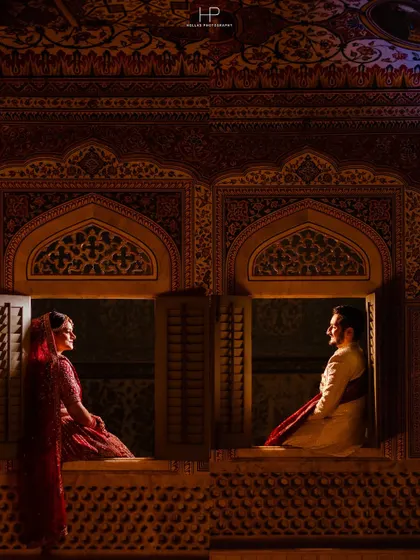 A cinematic shot of a couple in traditional attire, framed by the ornate windows of a palace. This style of photography uses architectural elements and dramatic lighting to create a regal and timeless portrait.