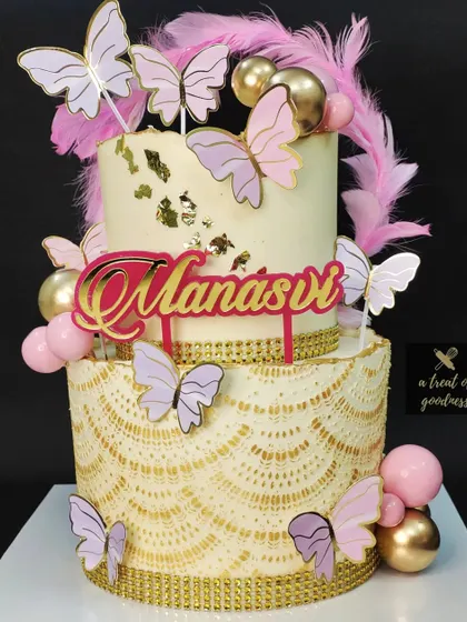 A beautiful two-tiered butterfly-themed cake. The bottom tier has a delicate lace-like stencil in gold, and the cake is covered in pink and purple butterflies and feathers.