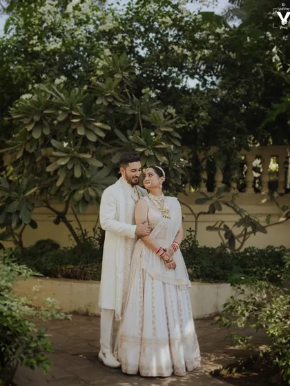 A classic portrait of the couple in matching white outfits, surrounded by lush greenery. Their elegant pose and the serene setting create a timeless and beautiful wedding photograph.