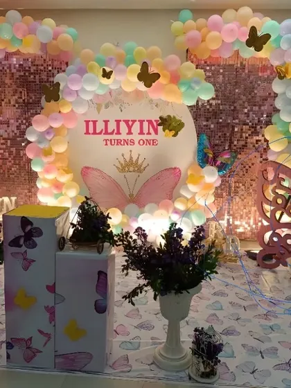 A butterfly-themed first birthday party with a pink shimmer wall. The setup features pastel-colored balloons, butterfly cutouts, and a custom-designed backdrop with the child's name.