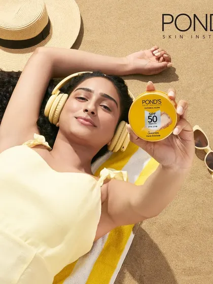 A lifestyle shot for a Pond's sunscreen powder campaign. The image portrays a relaxed, summery scene with a model on a beach towel, seamlessly integrating the product into a relatable, everyday moment.