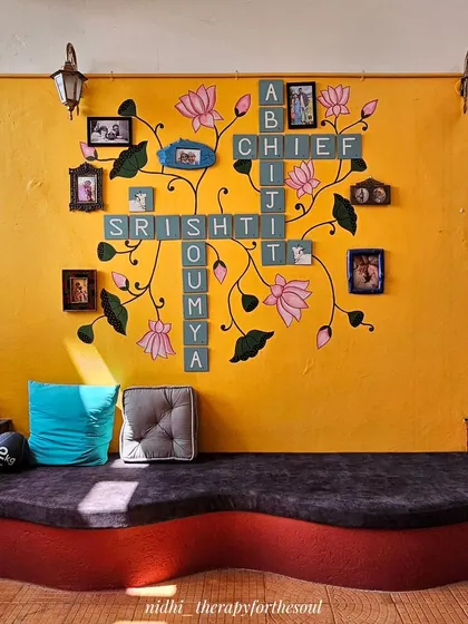 A full view of the custom Scrabble wall art, where family names are interwoven with hand-painted Pichwai lotus vines against a vibrant yellow wall.