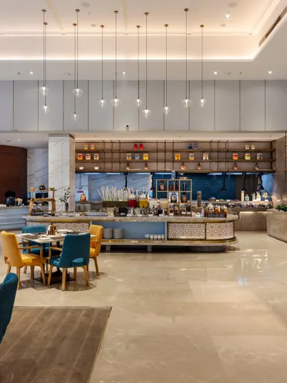 The all-day dining area features a central island buffet and an open show kitchen to create an engaging environment. The high ceilings and pendant lighting enhance the sense of space, while varied seating caters to different group sizes.