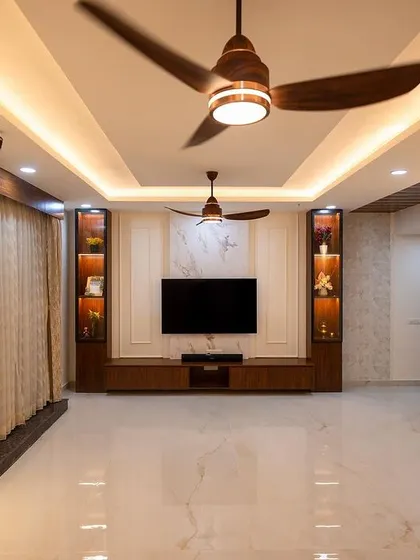A wide view of a spacious and elegant living room. The design features a sophisticated TV unit with display shelves, beautiful false ceiling with dual fans, and rich wooden accents for a timeless appeal.