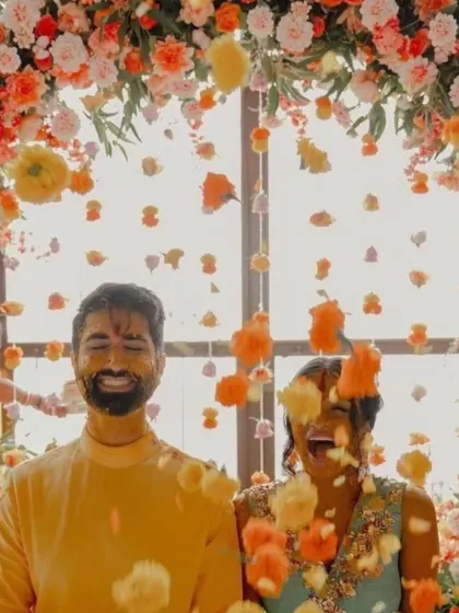 A candid shot of a couple enjoying a flower shower during their Haldi ceremony, framed by a vibrant floral arch.
