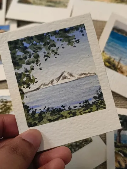 Peeking through lush green foliage to a view of a mountain across the water. This hand-painted Polaroid uses framing to create depth and draw your eye into the landscape.